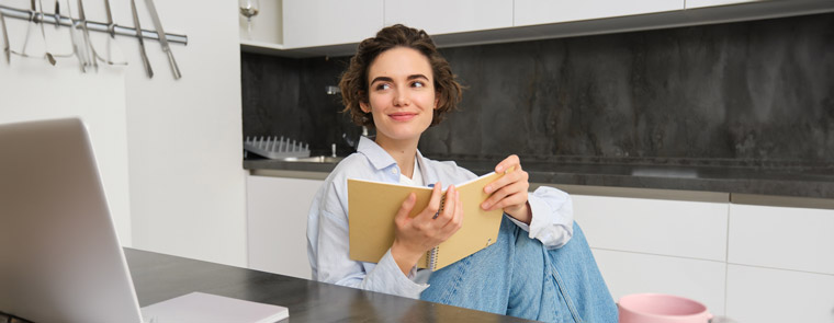 young-woman-sitting-kitchen-reading-her-notes-notebook-smiling-girl-home-doing-homework